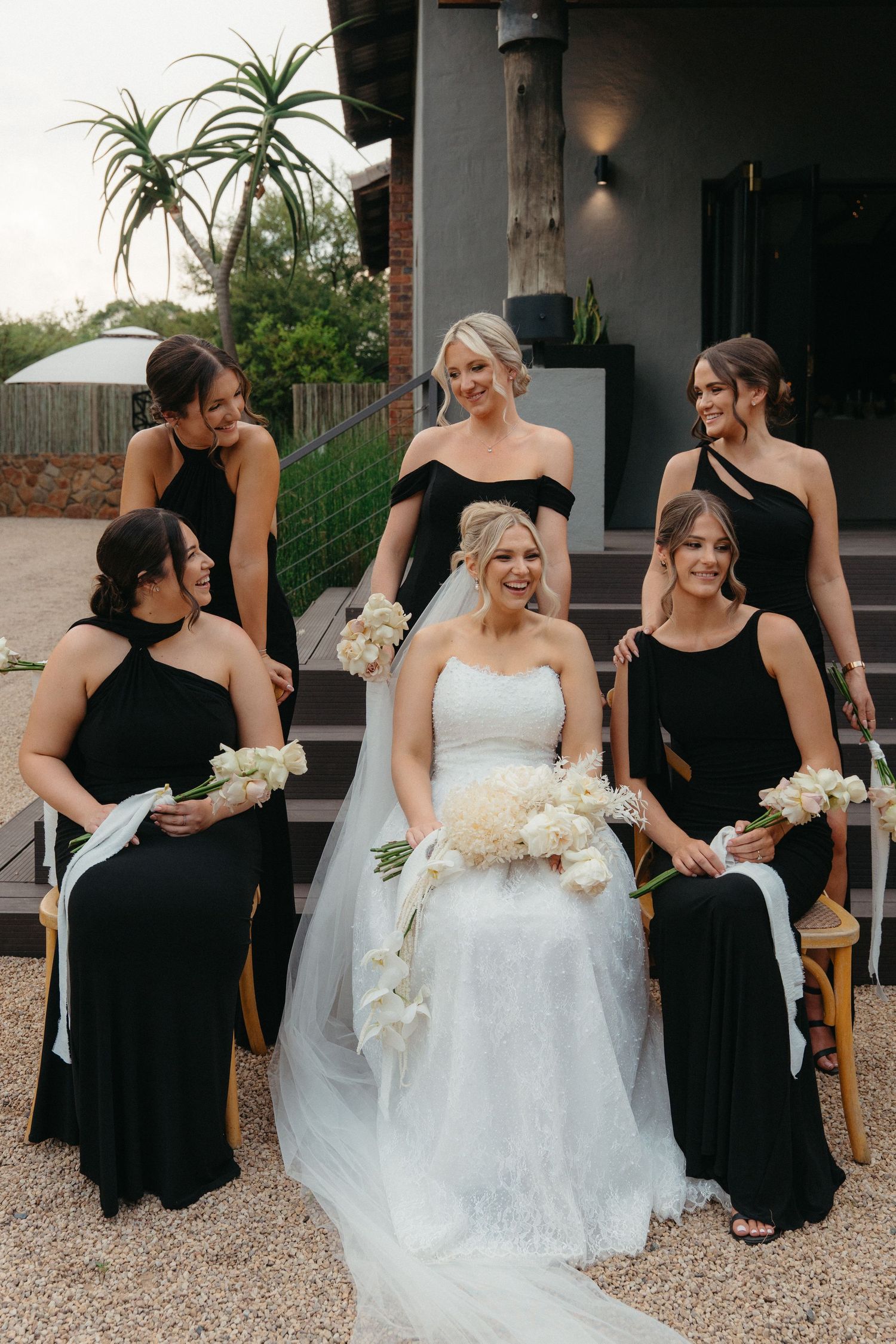 Wedding party in elegant black bridesmaid dresses poses with bride in white gown holding floral bouquets.