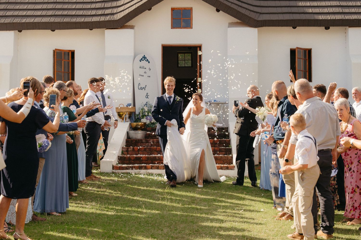 Wedding guests gather outside a rustic building for a celebratory reception on a sunny afternoon.
