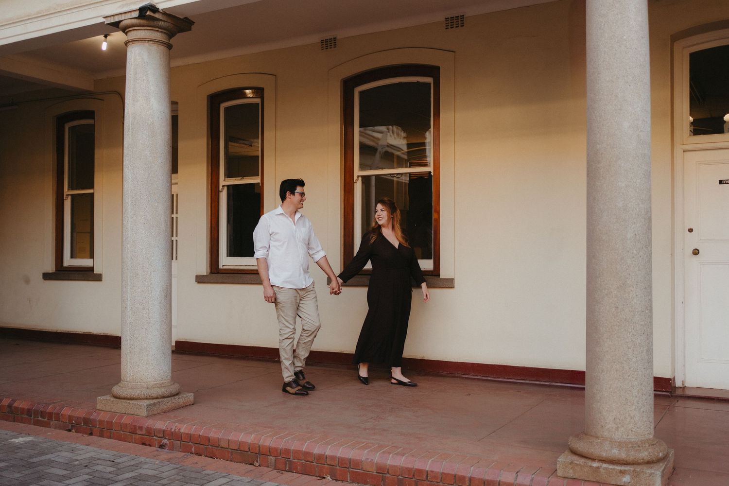 Couple walking hand in hand along a covered veranda of a classic Pretoria heritage building.