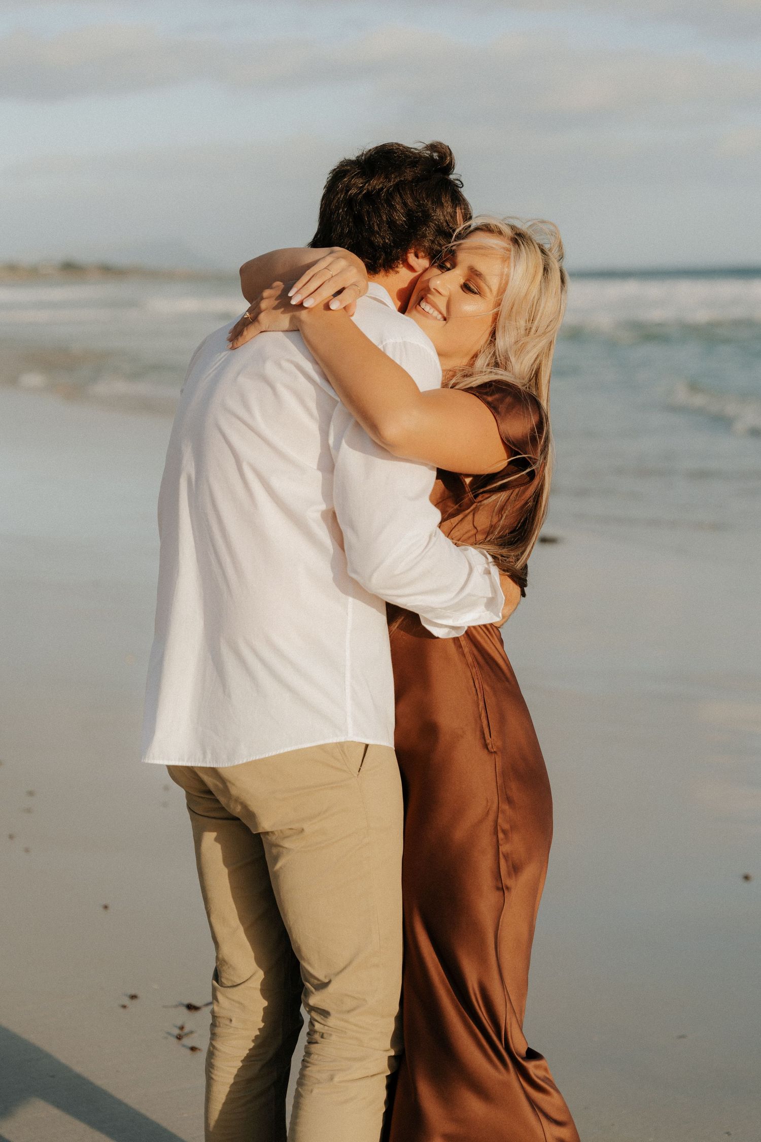 A couple embraces romantically on a sandy beach, woman in brown satin dress smiling joyfully.
