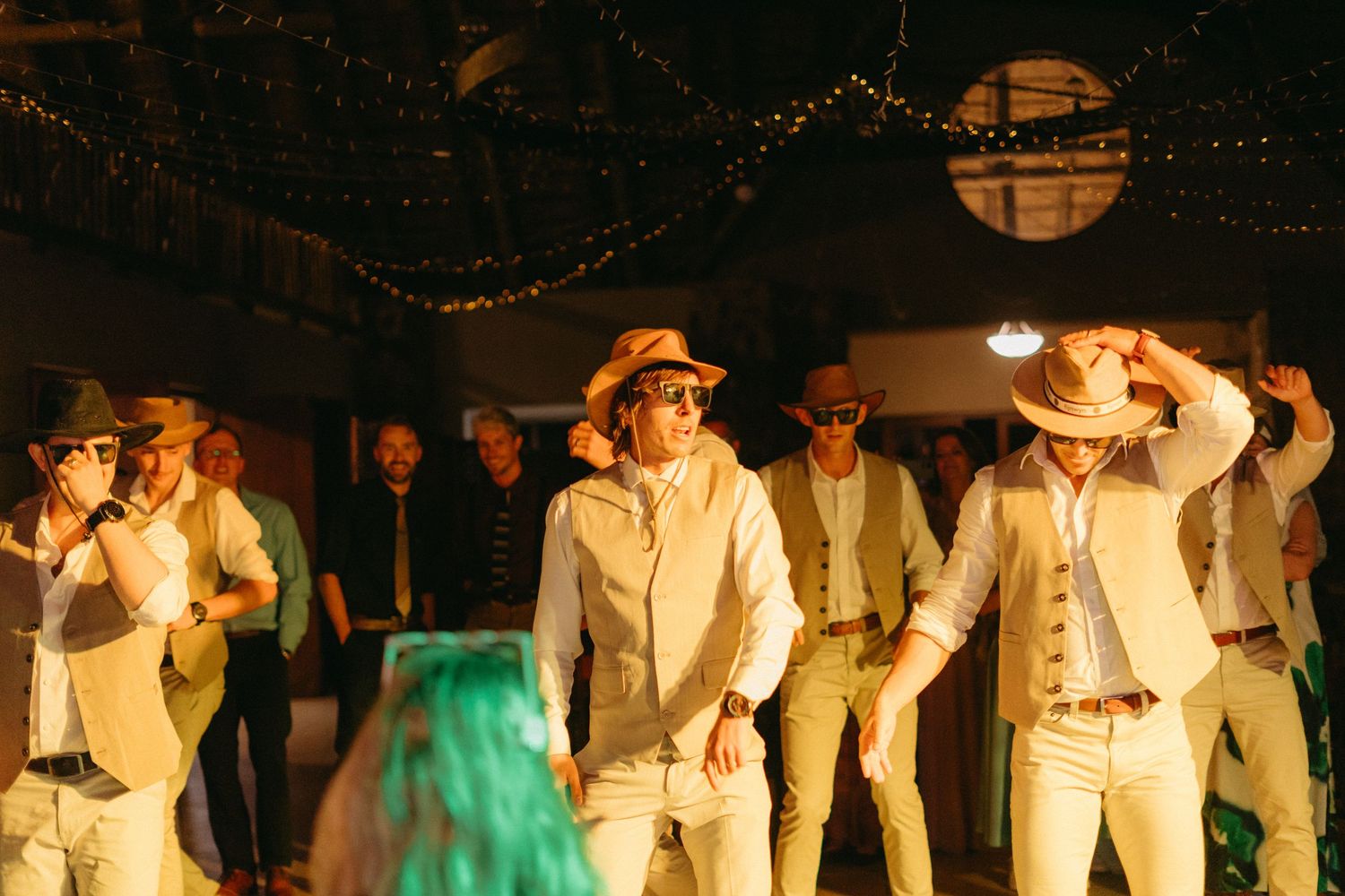 Wedding party in white attire dancing under warm lighting at evening reception.