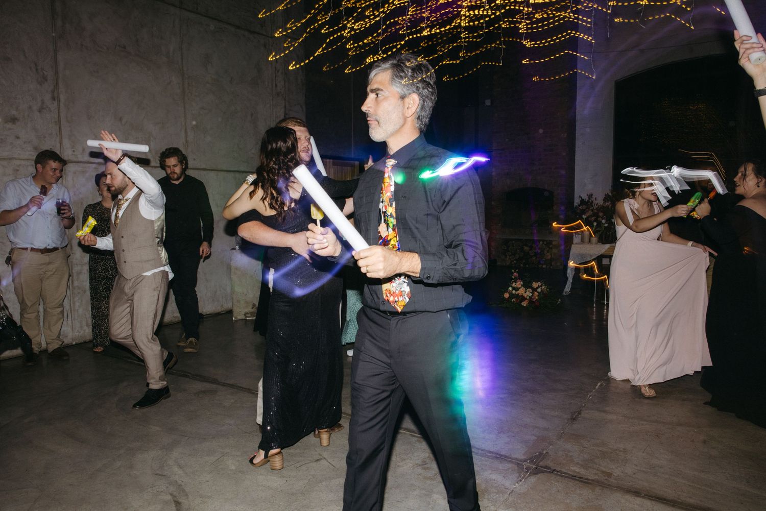 Wedding guests dance under colorful lights during an evening reception celebration.