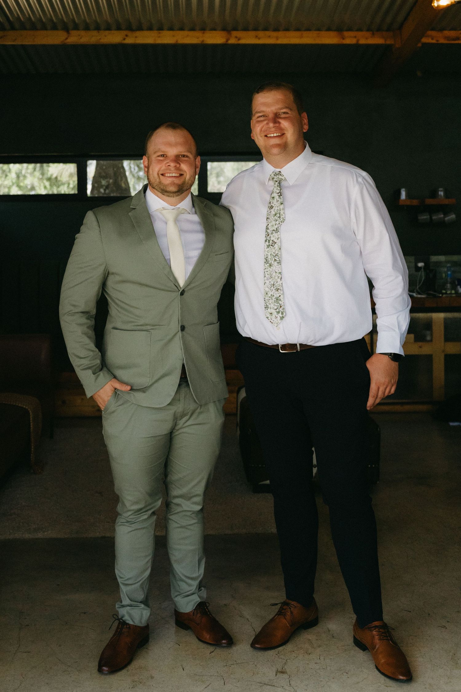 Two men in formal attire smile together under a covered outdoor space.
