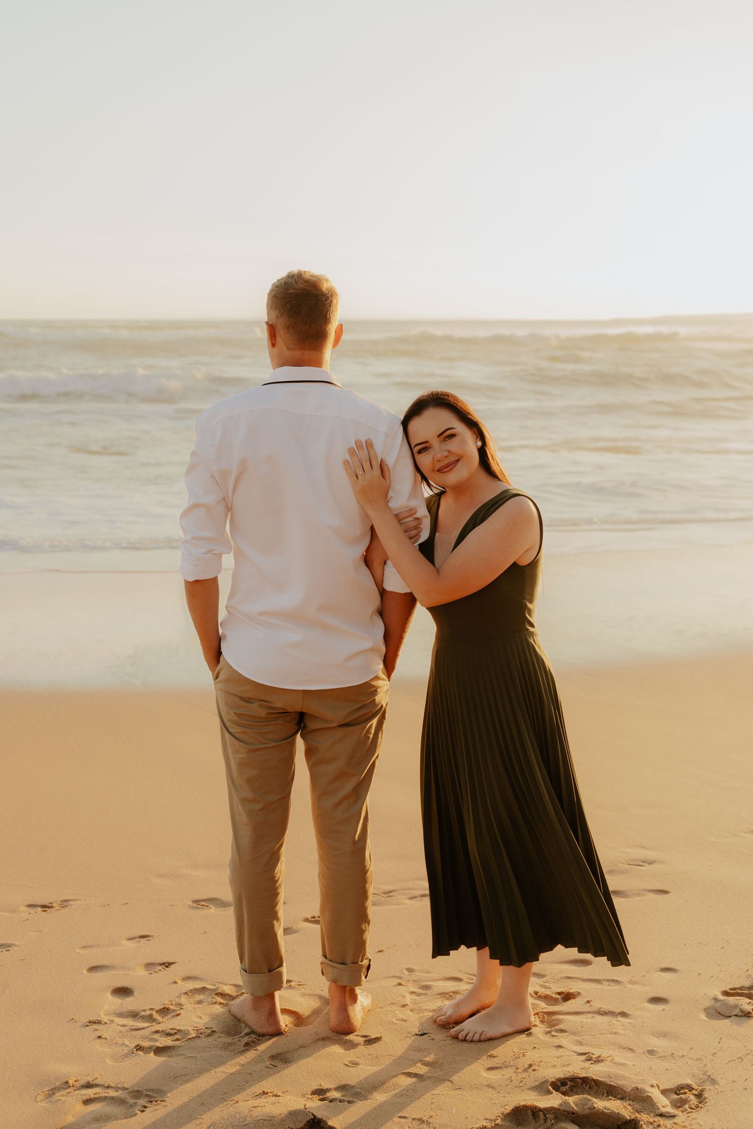 A couple stands barefoot on a sandy beach at sunset in a Hartenbos engagement shoot