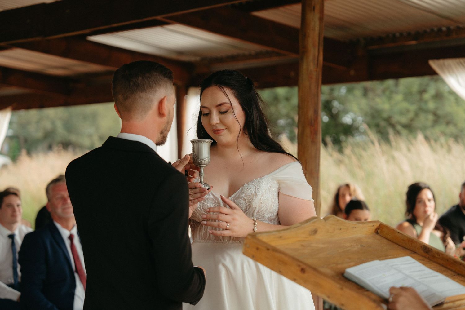 A couple exchanges rings during their wedding ceremony under a rustic wooden pavilion.