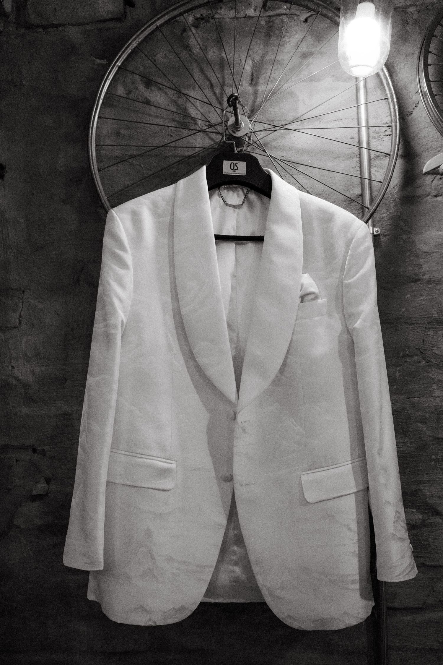 A crisp white formal tuxedo jacket hanging against a rustic wall with a bicycle wheel backdrop.