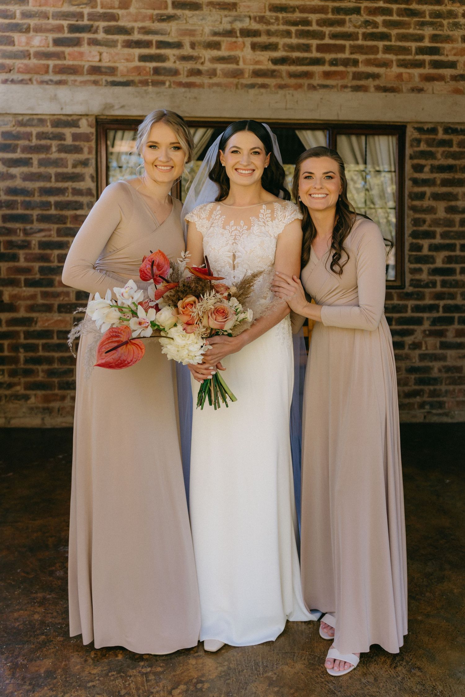 Bridesmaids in blush pink dresses pose with bride in white gown against brick wall.