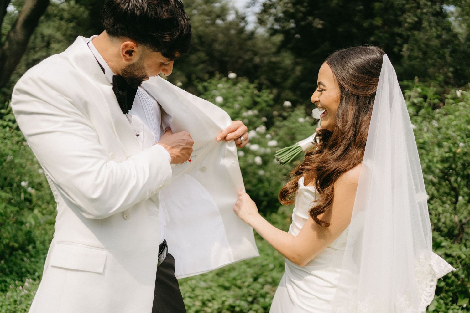 A couple in white wedding attire laughing together during a fun interaction in a lush garden setting.