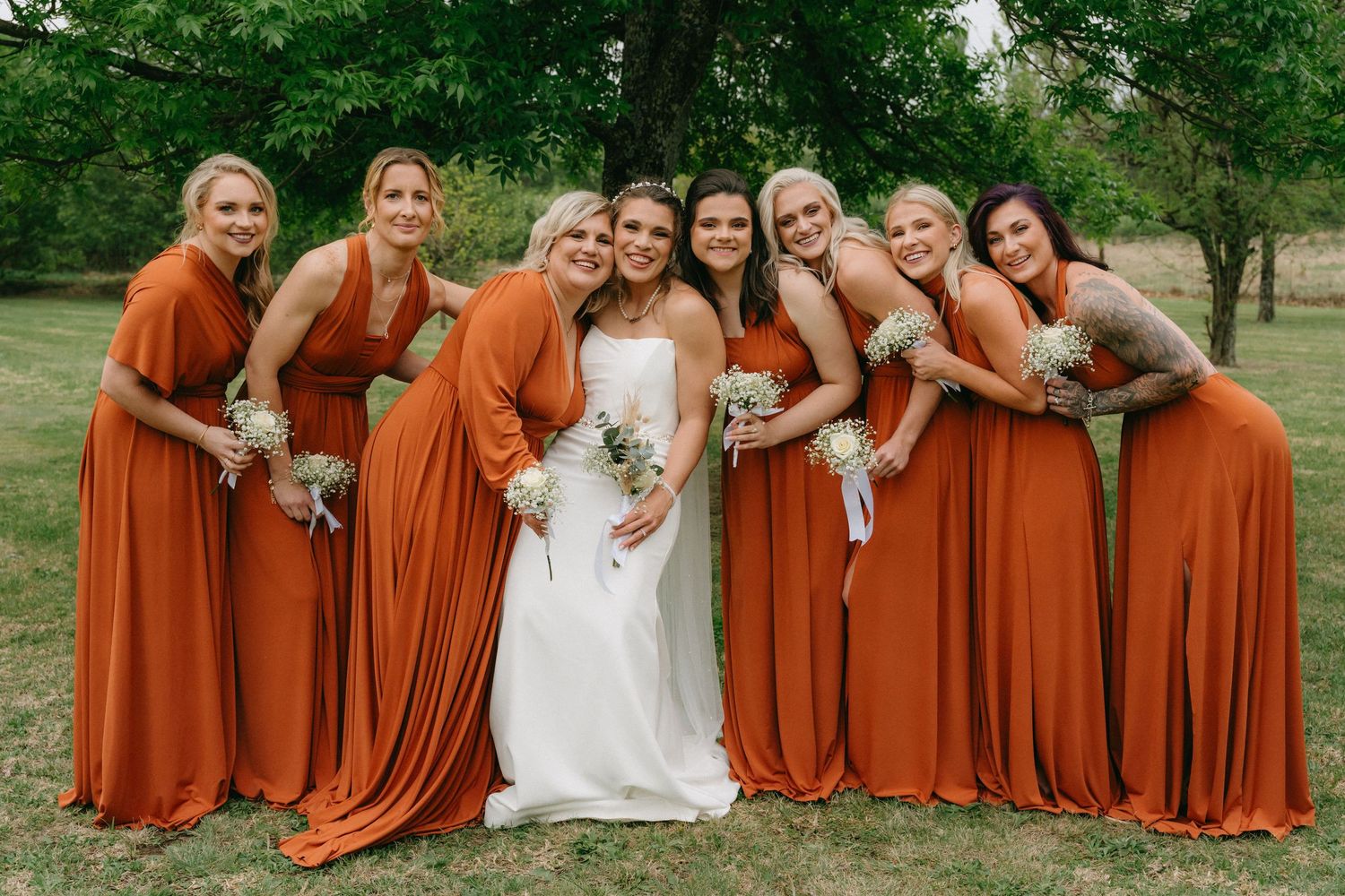 Bridal party in rust-colored dresses poses together on grassy lawn.