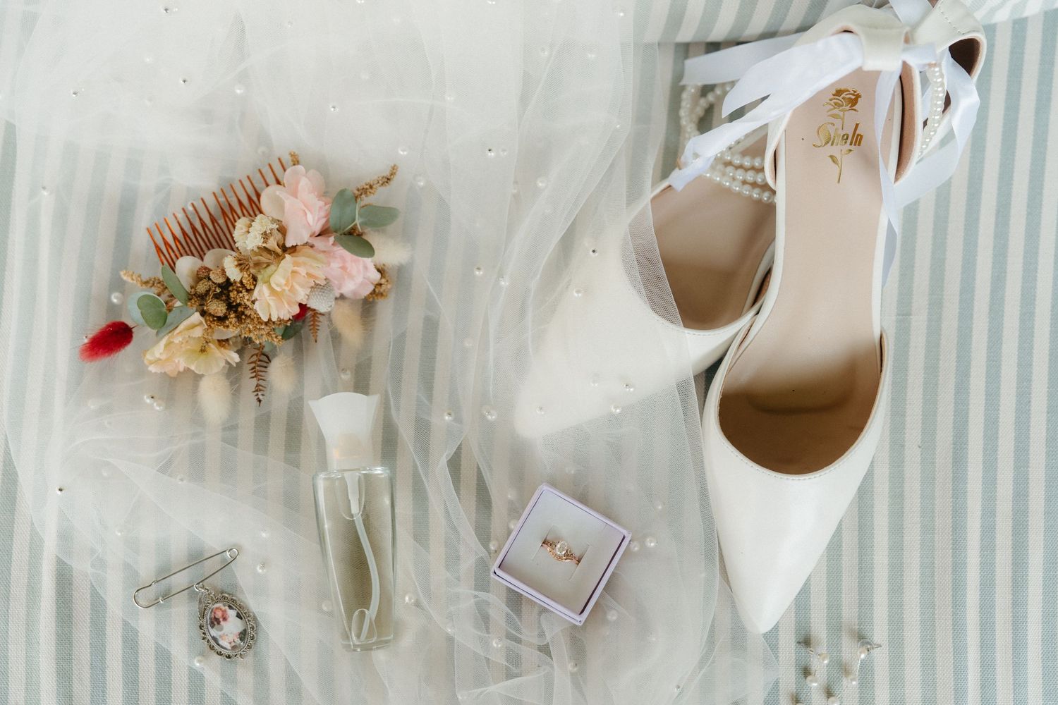 Wedding details flatlay with white heels, floral hair comb, ring box and champagne glass.