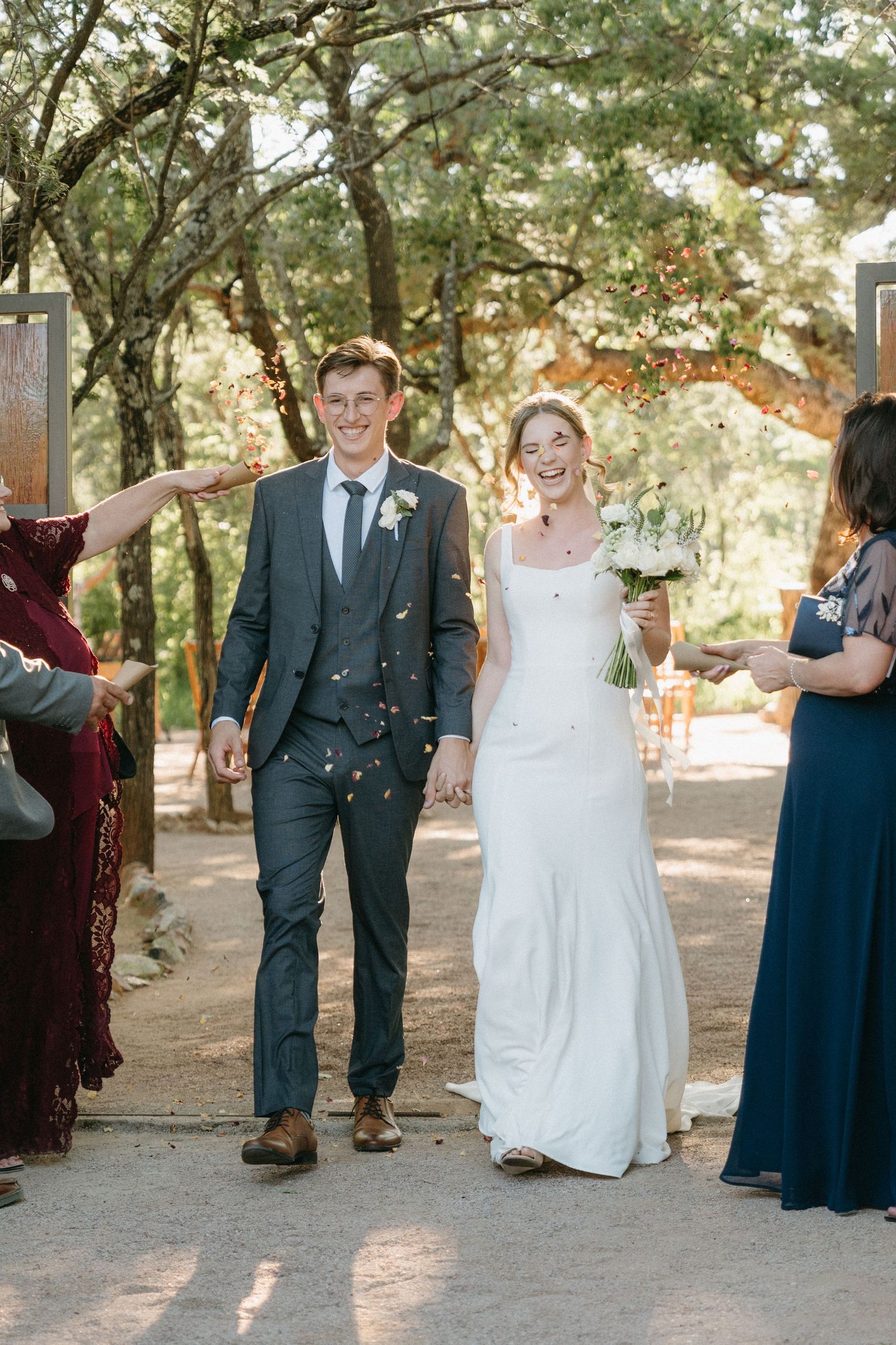 Guests throw confetti at a newlywed couple walking along an outdoor path during their wedding celebration.