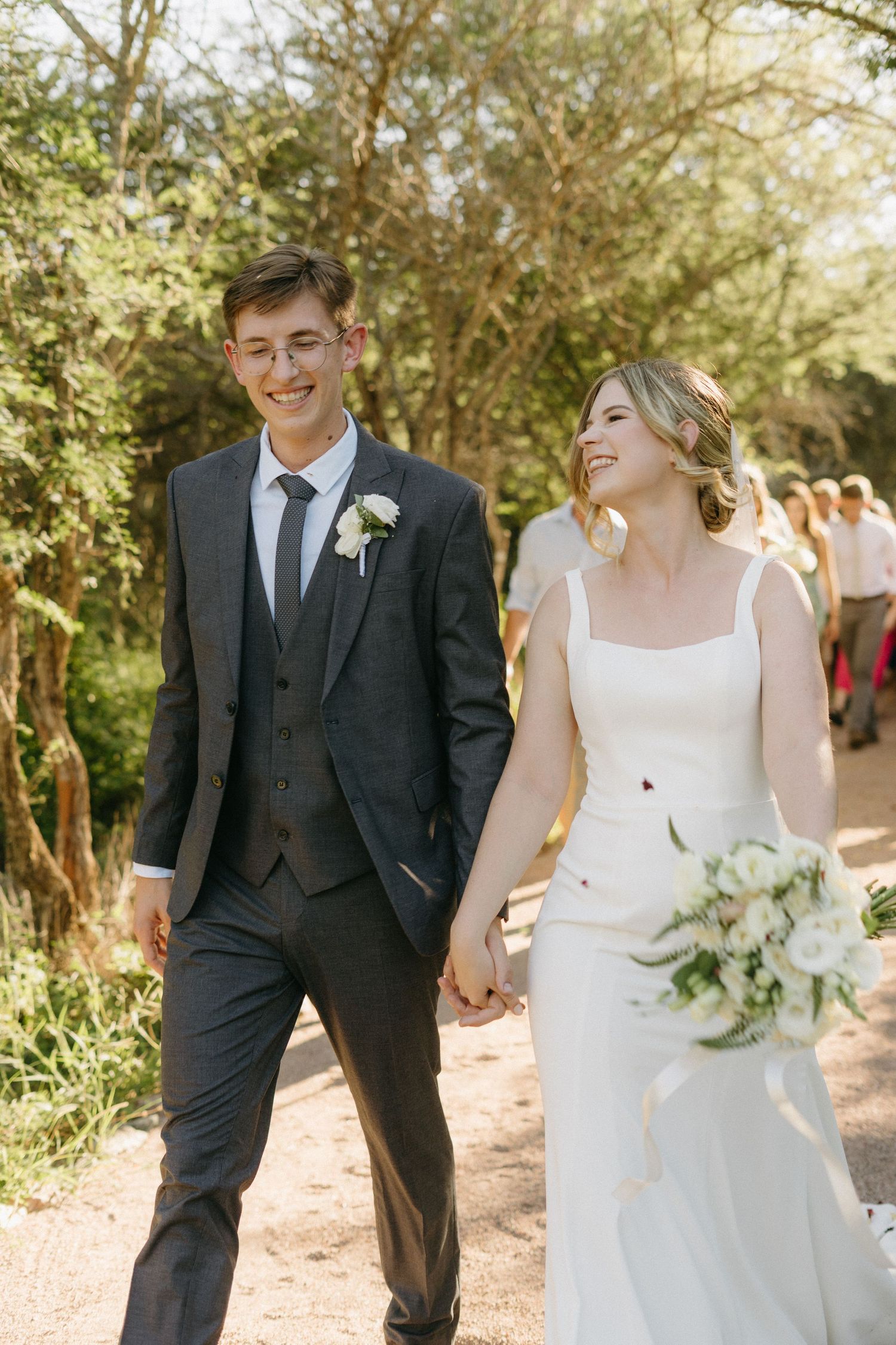 A happy couple walks along an outdoor path on their wedding day, wearing elegant formal attire.