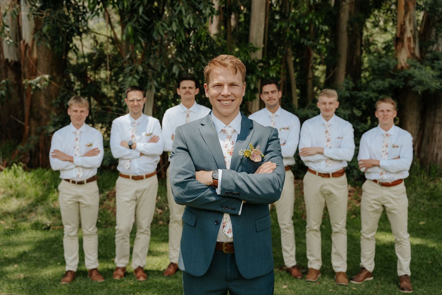 A wedding party poses outdoors with groomsmen wearing light khaki pants and blue dress shirts against a natural backdrop.