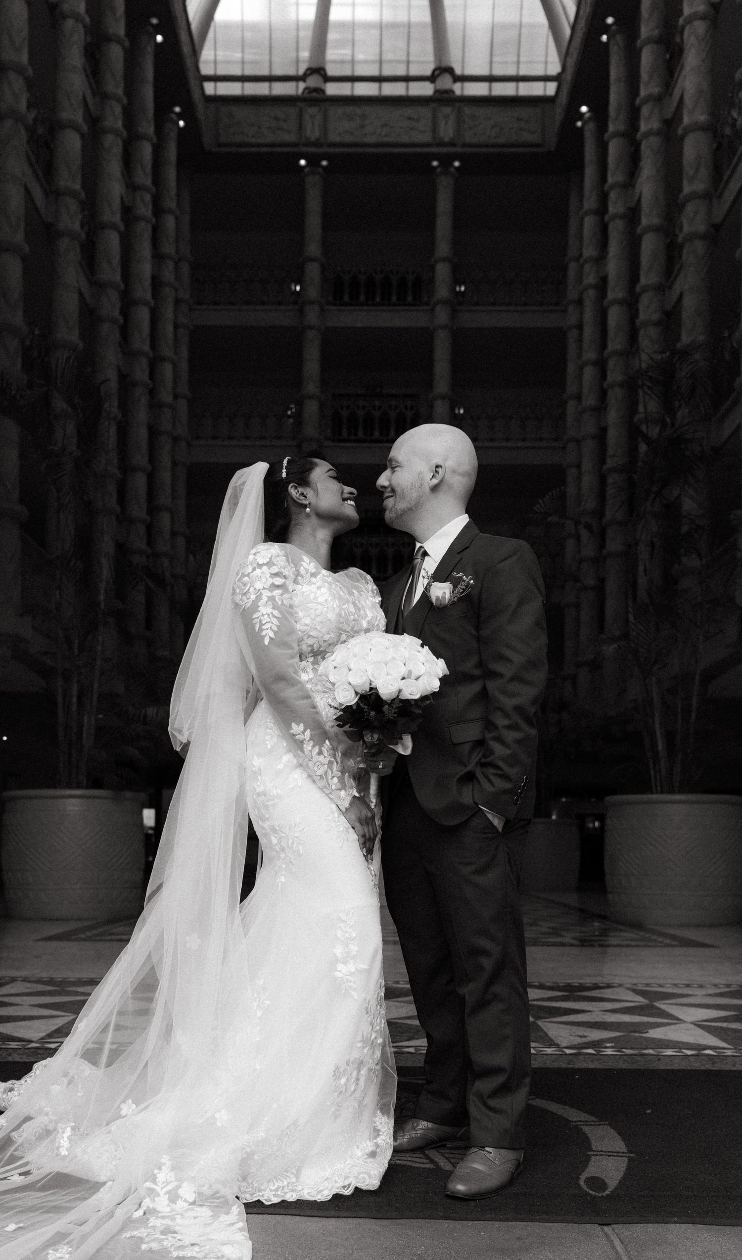 Wedding couple in elegant venue with columns stands close together as bride holds white bouquet in black and white photo.