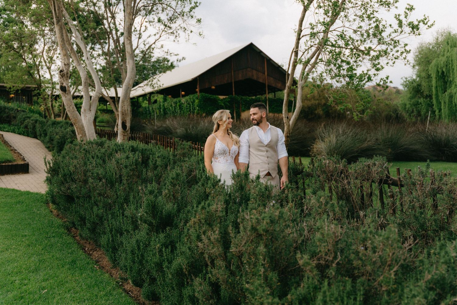 A couple stands together in a garden pathway surrounded by lush greenery and trees.