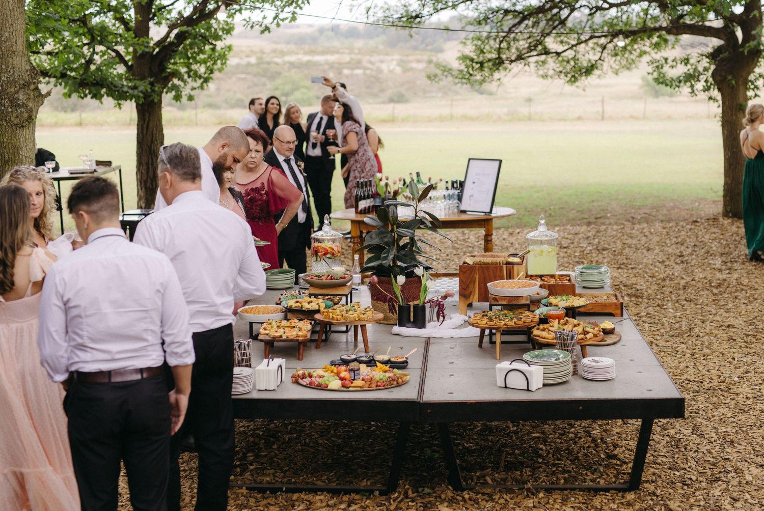 Wedding guests gather around an outdoor buffet table filled with food and catering displays under trees.