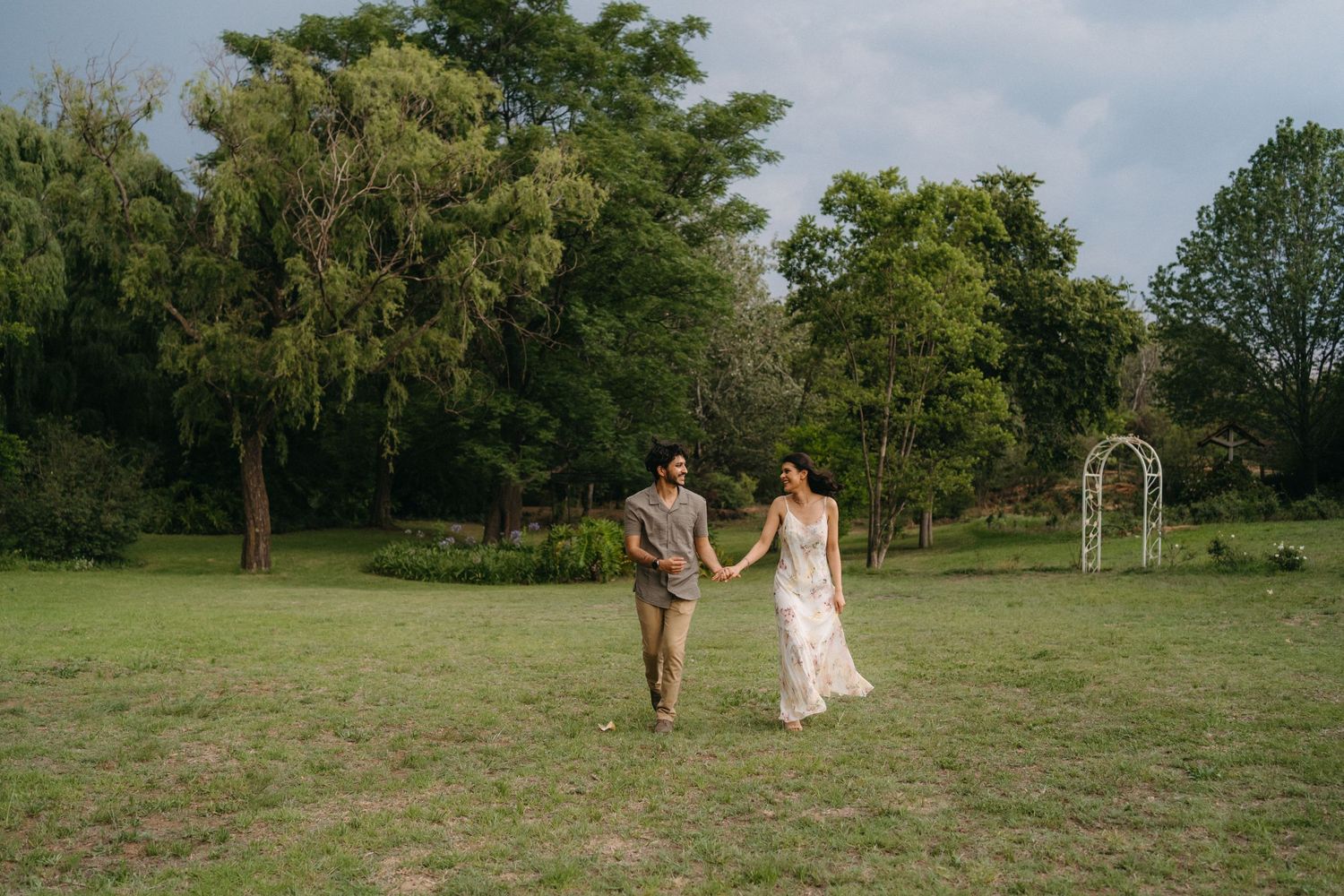 Couple running hand in hand through an open field with garden arch in background.