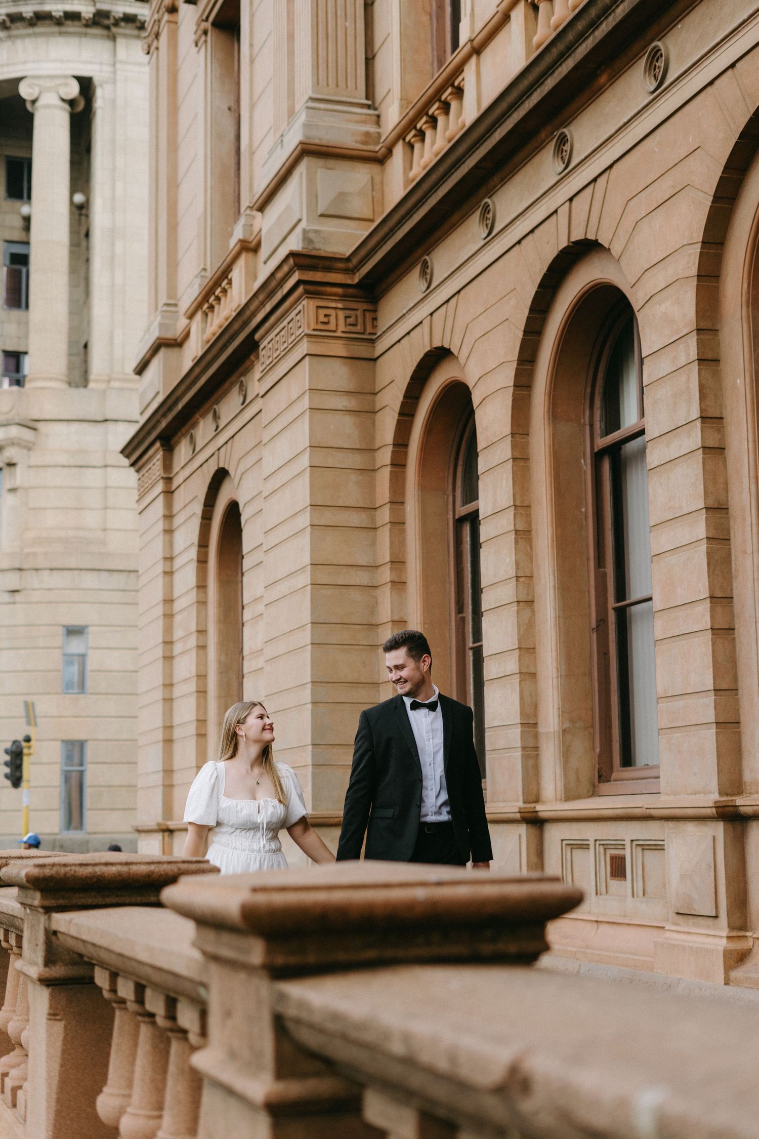 A romantic couple in formal attire stands on a grand historic building's balcony with ornate columns and architecture.