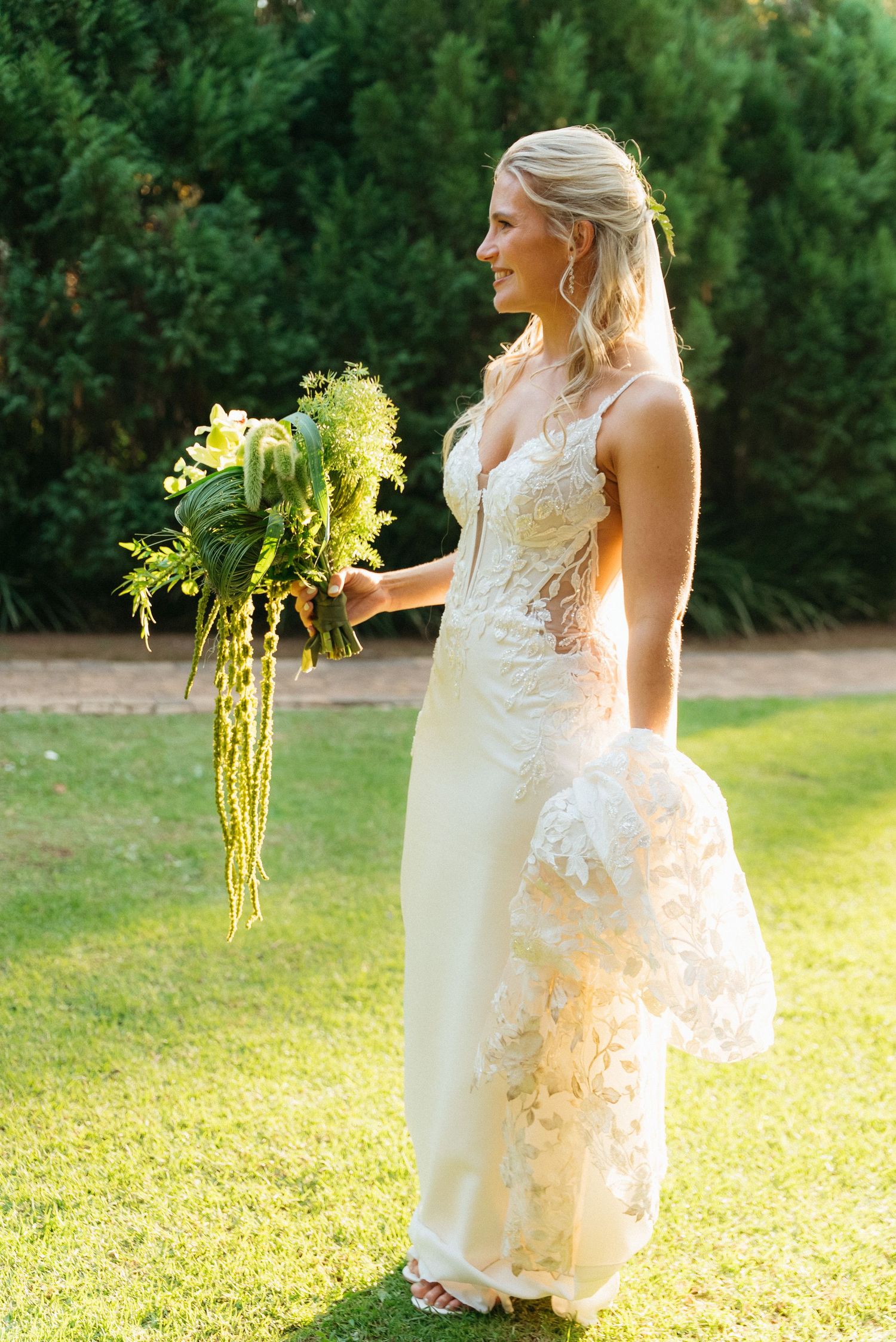 Blonde bride in lace wedding gown holding green tropical bouquet standing on grass with trees behind.