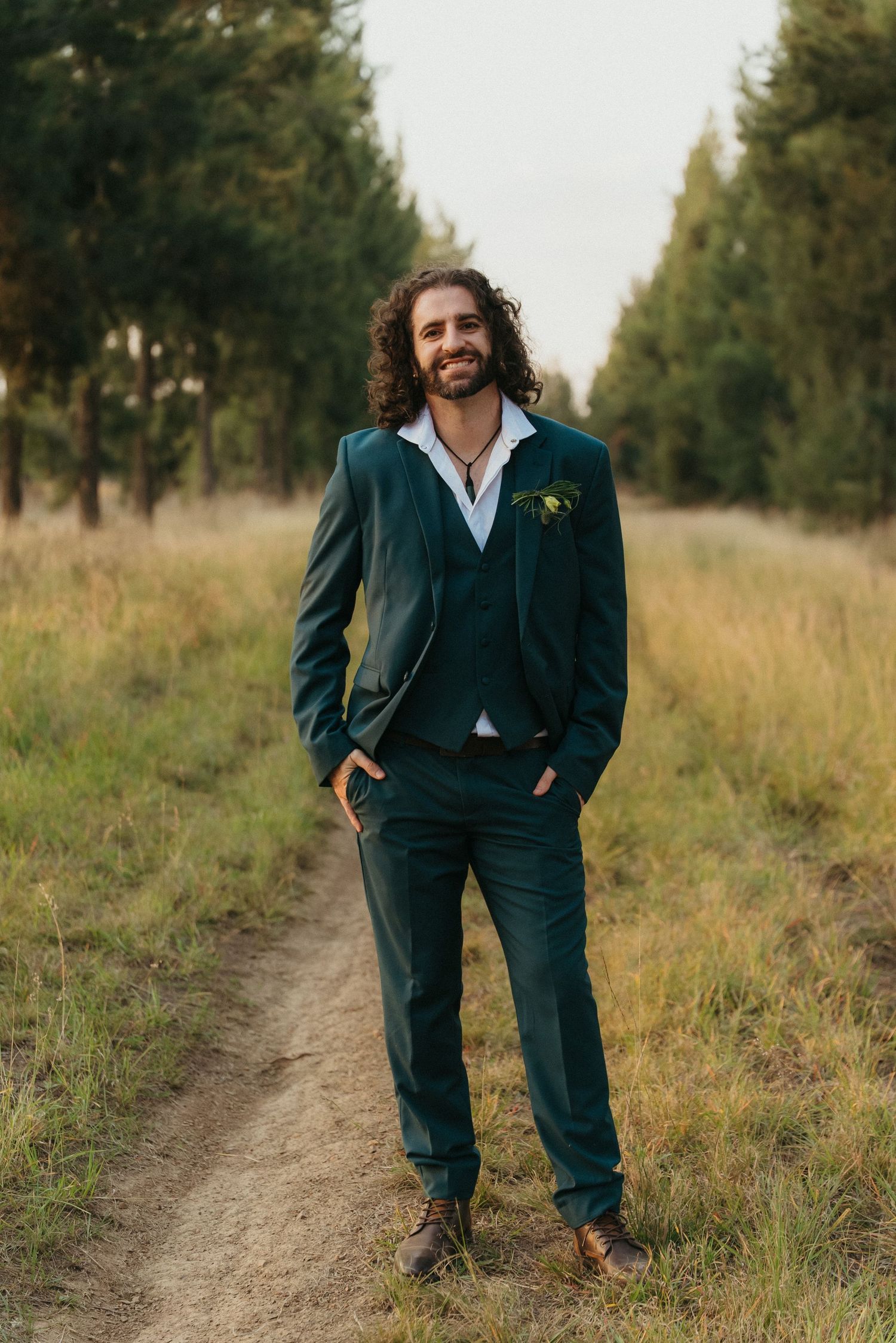 Groom in dark green three-piece suit with boutonniere stands on dirt path surrounded by tall trees.