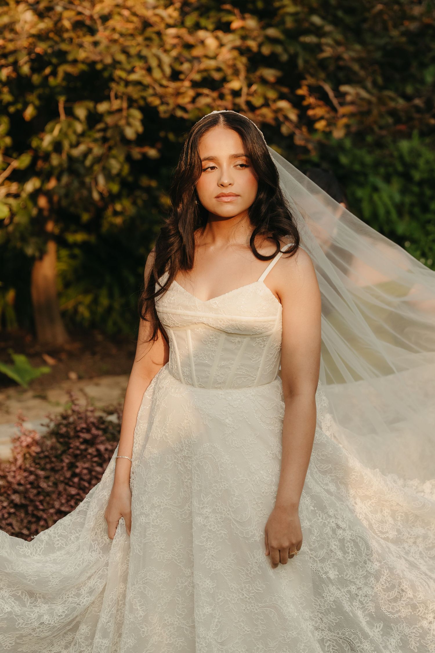 Bride in lace wedding gown and flowing veil poses outdoors amid golden autumn foliage in soft warm light.