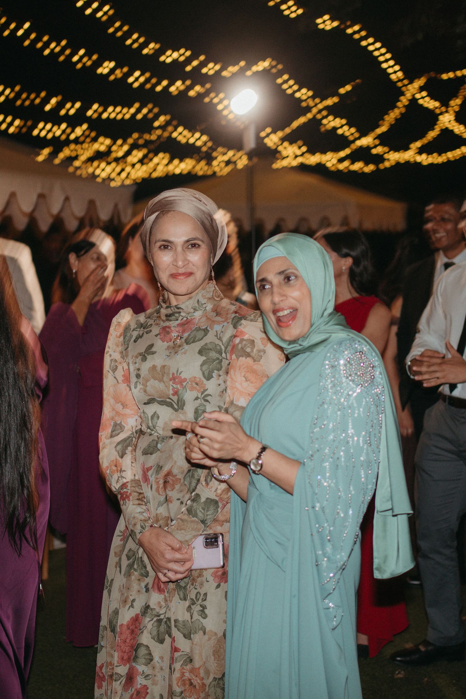 Two women in elegant attire smile together at a festive evening event with warm string lights overhead.
