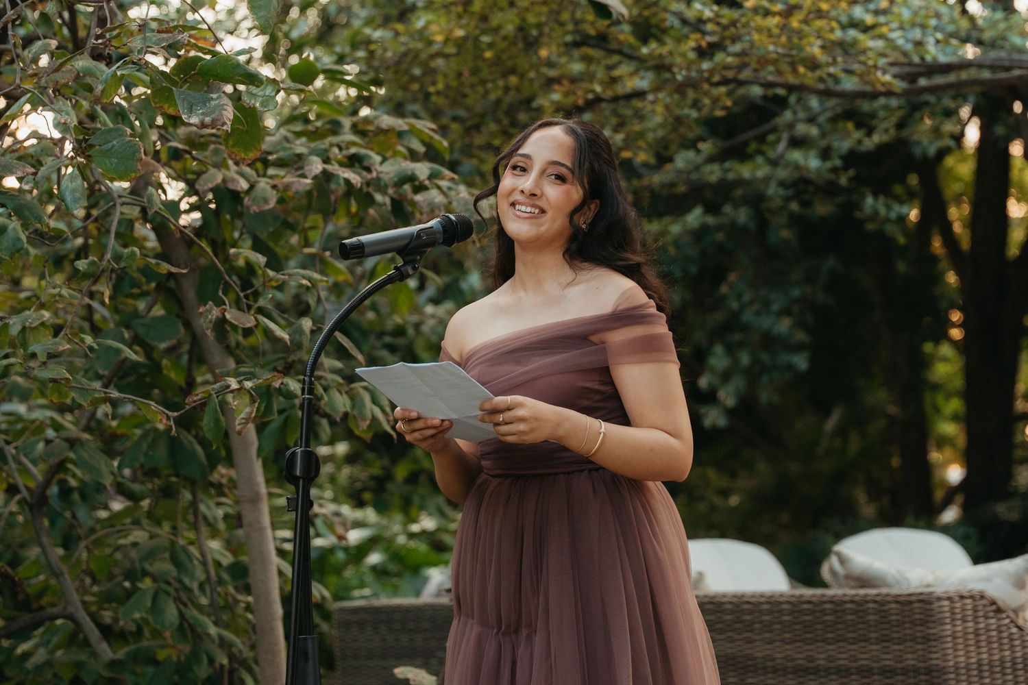 Woman in mauve off-shoulder tulle gown speaking at outdoor podium with microphone amid lush greenery.