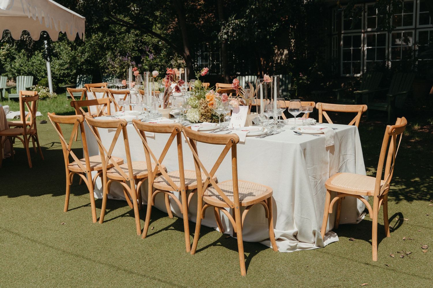 Elegant outdoor wedding reception at Beechwood Gardens with wooden cross-back chairs, white linen, floral centerpieces on a green lawn.