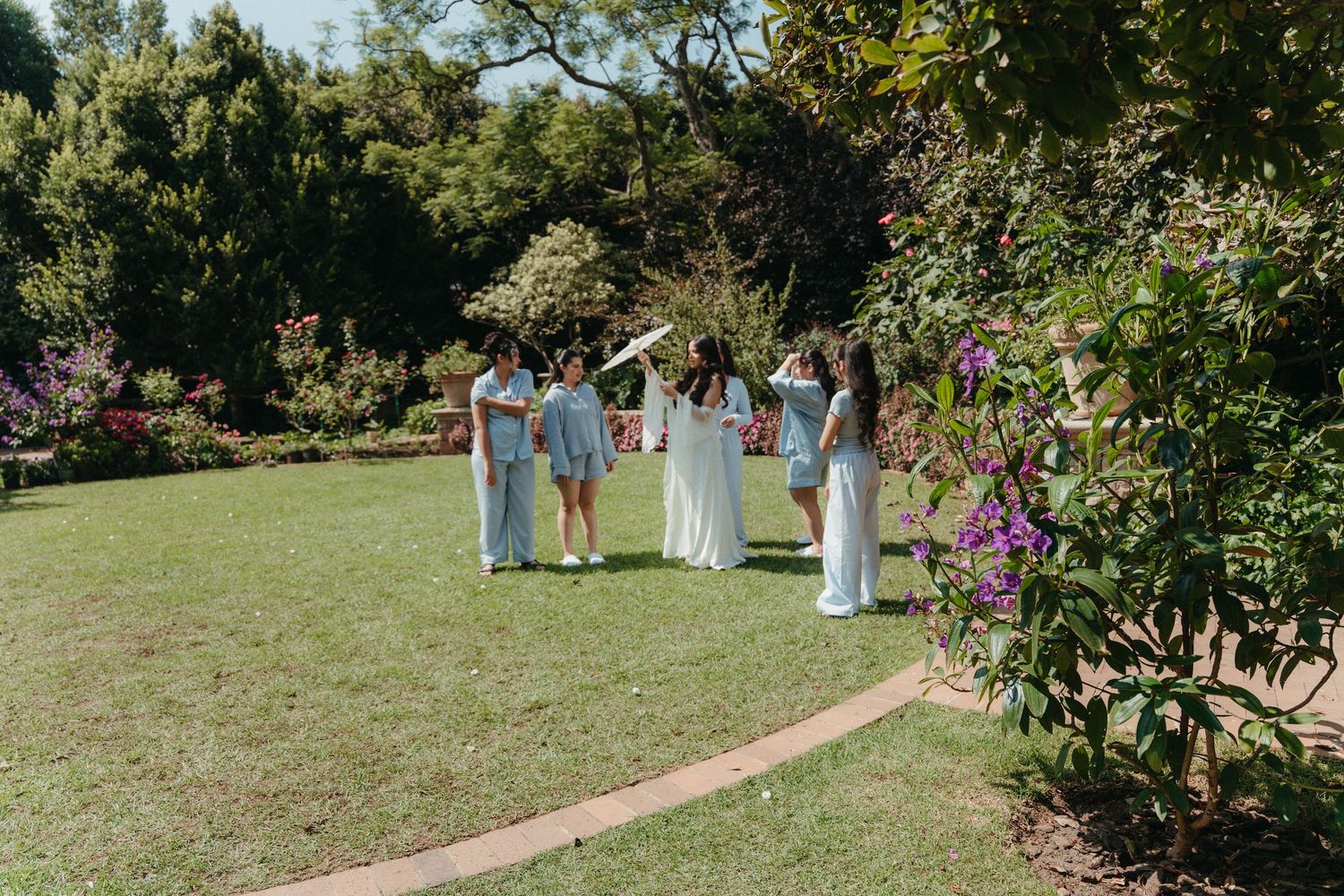 Bridesmaids in light blue dresses stand with bride in white gown on lush green garden lawn surrounded by colorful flowering shrubs.