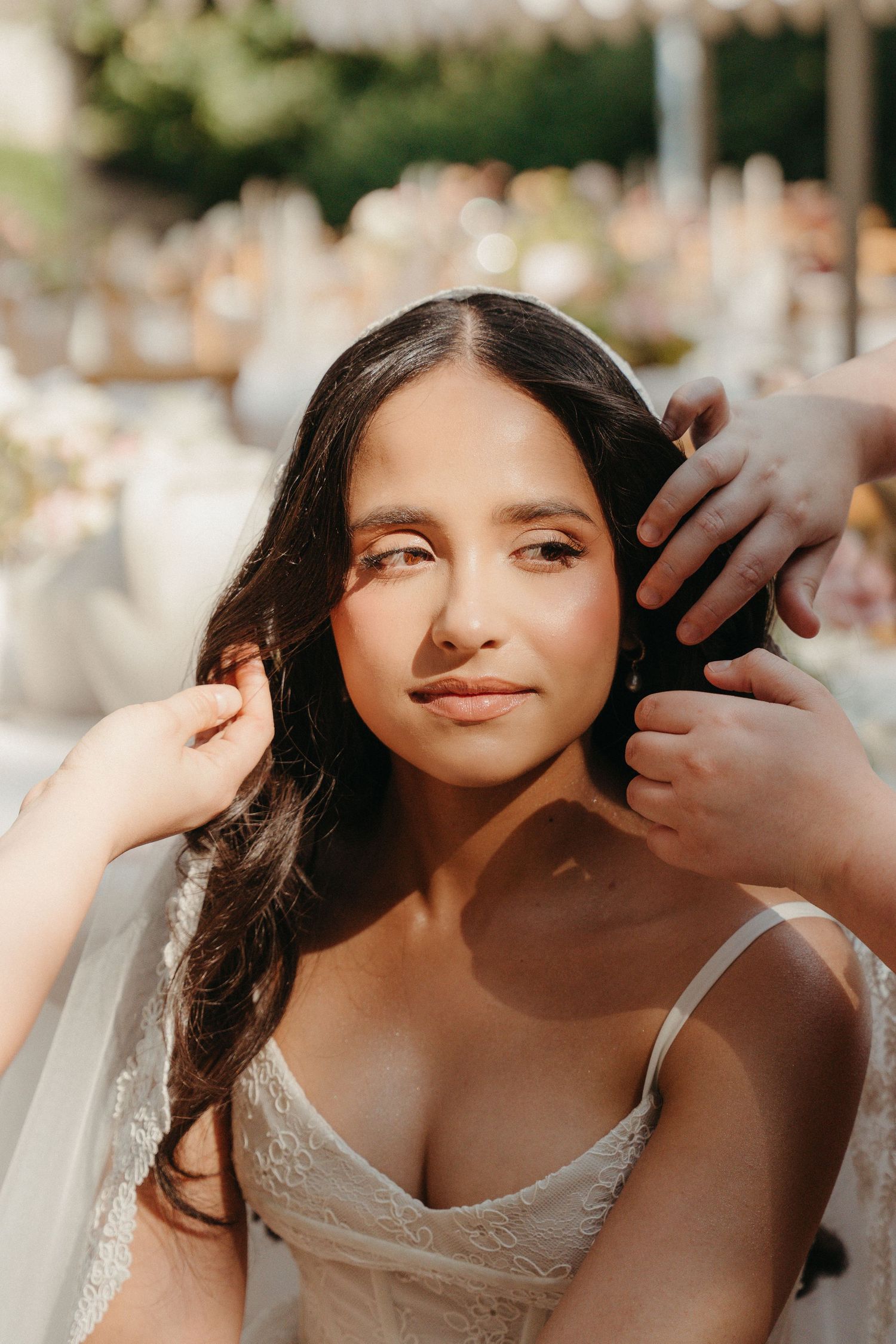 A bride in a white lace dress gets her veil adjusted outdoors at a wedding reception in soft golden light.