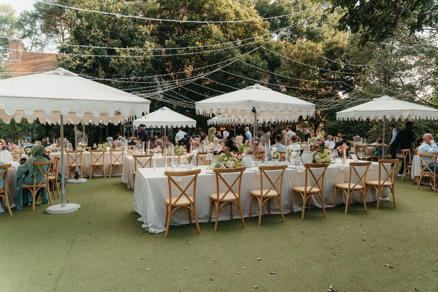 Elegant outdoor wedding reception with white market umbrellas, wooden cross-back chairs, and floral centerpieces on a green lawn.
