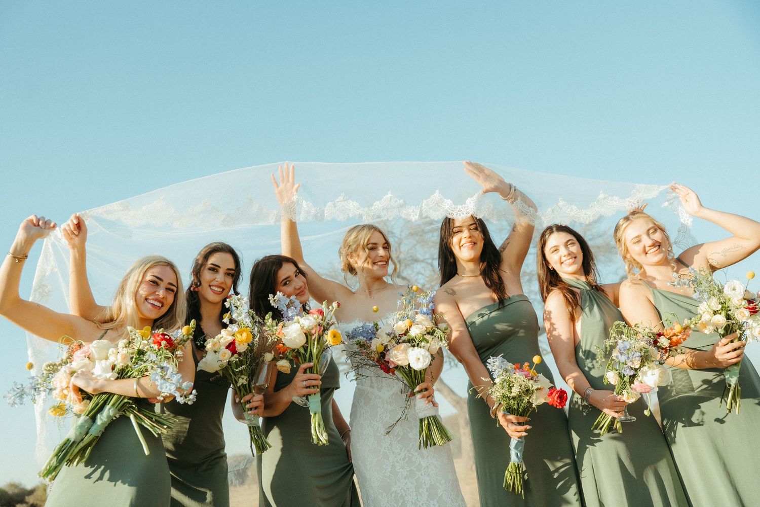 Bride and bridesmaids in sage green dresses holding colorful bouquets under a flowing veil against a Limpopo blue sky.