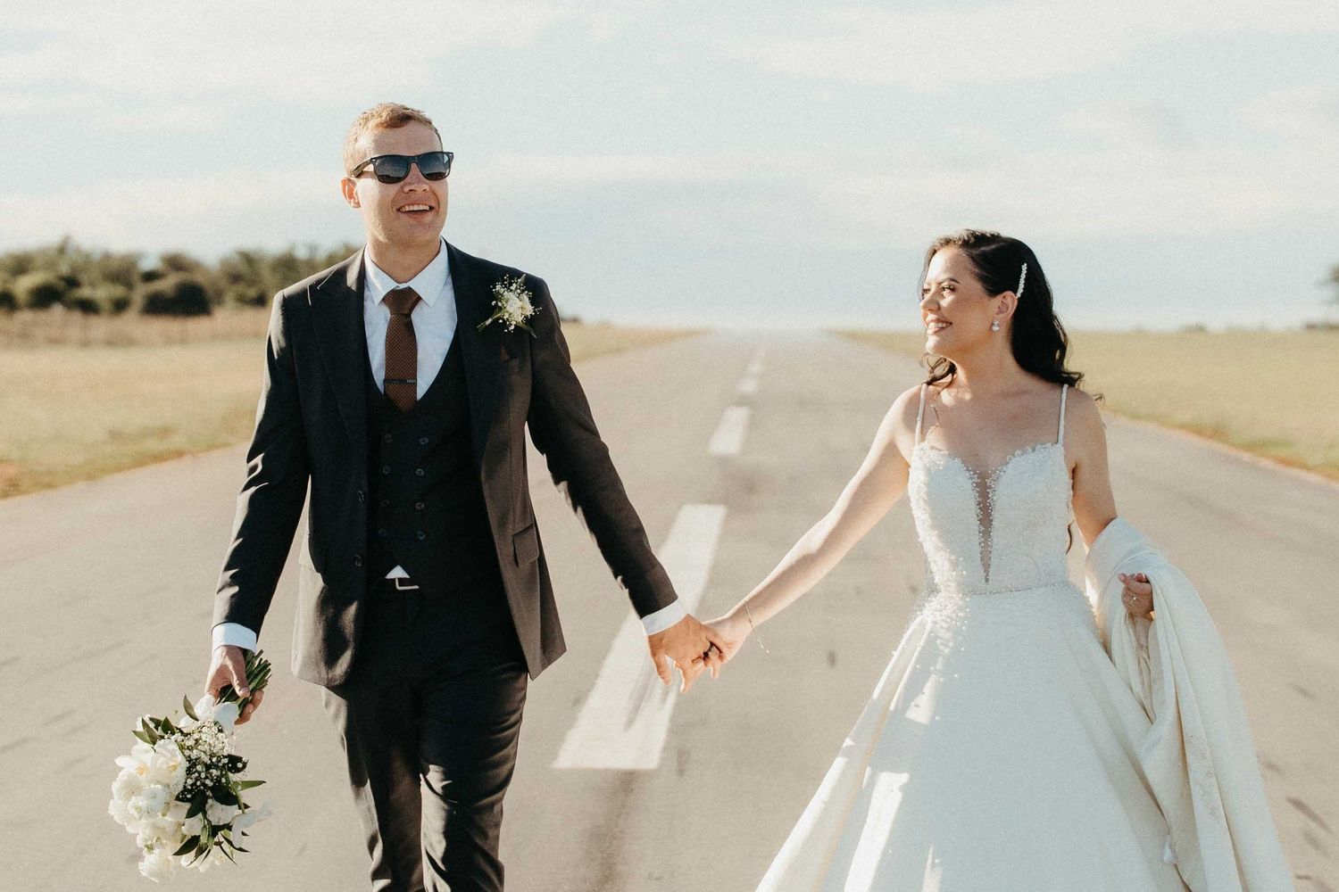 Bride and groom walk hand-in-hand on a runway, smiling, groom in dark suit with sunglasses, bride in white ballgown.
