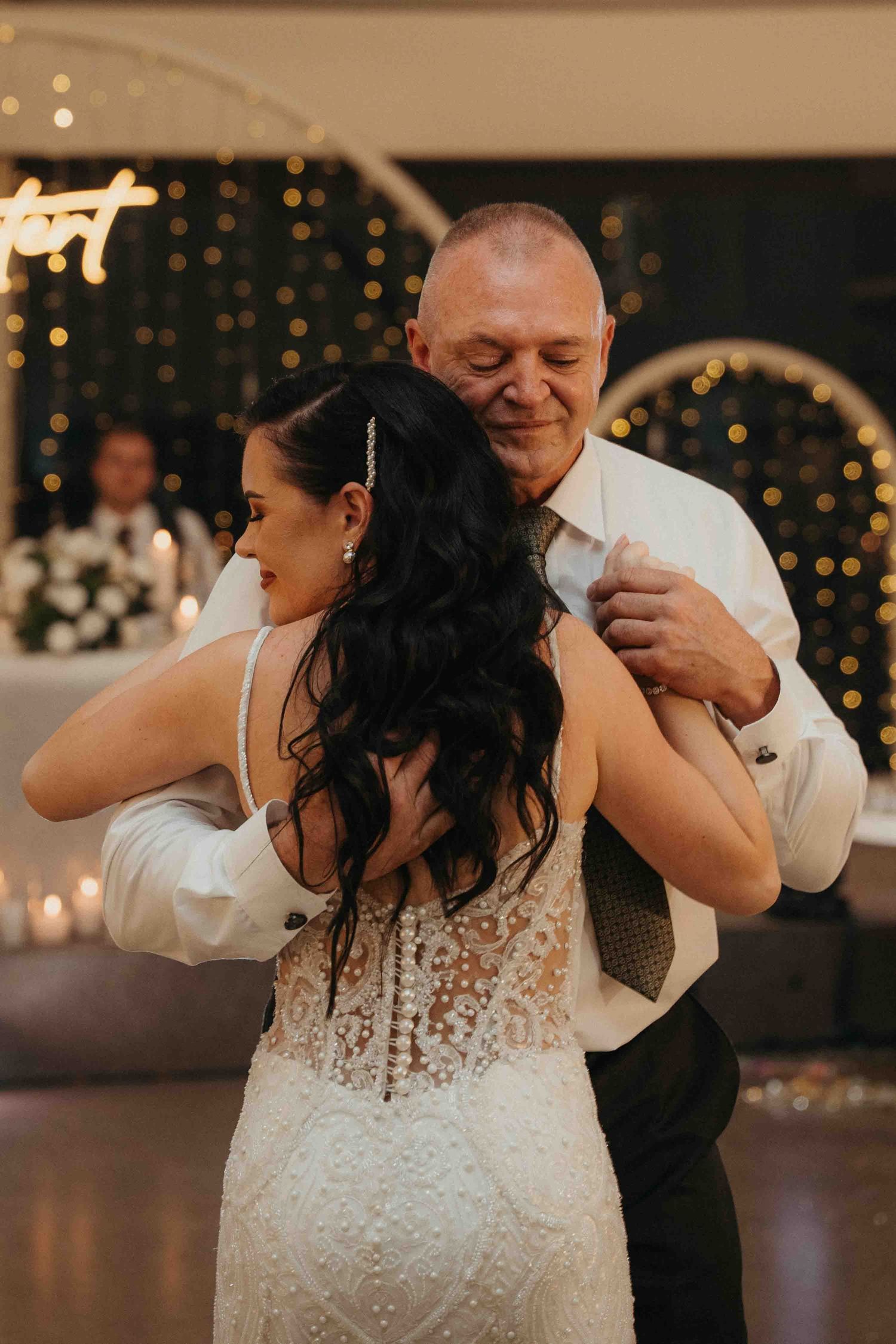 Bride and father share an emotional first dance at wedding reception with warm bokeh lights in background.