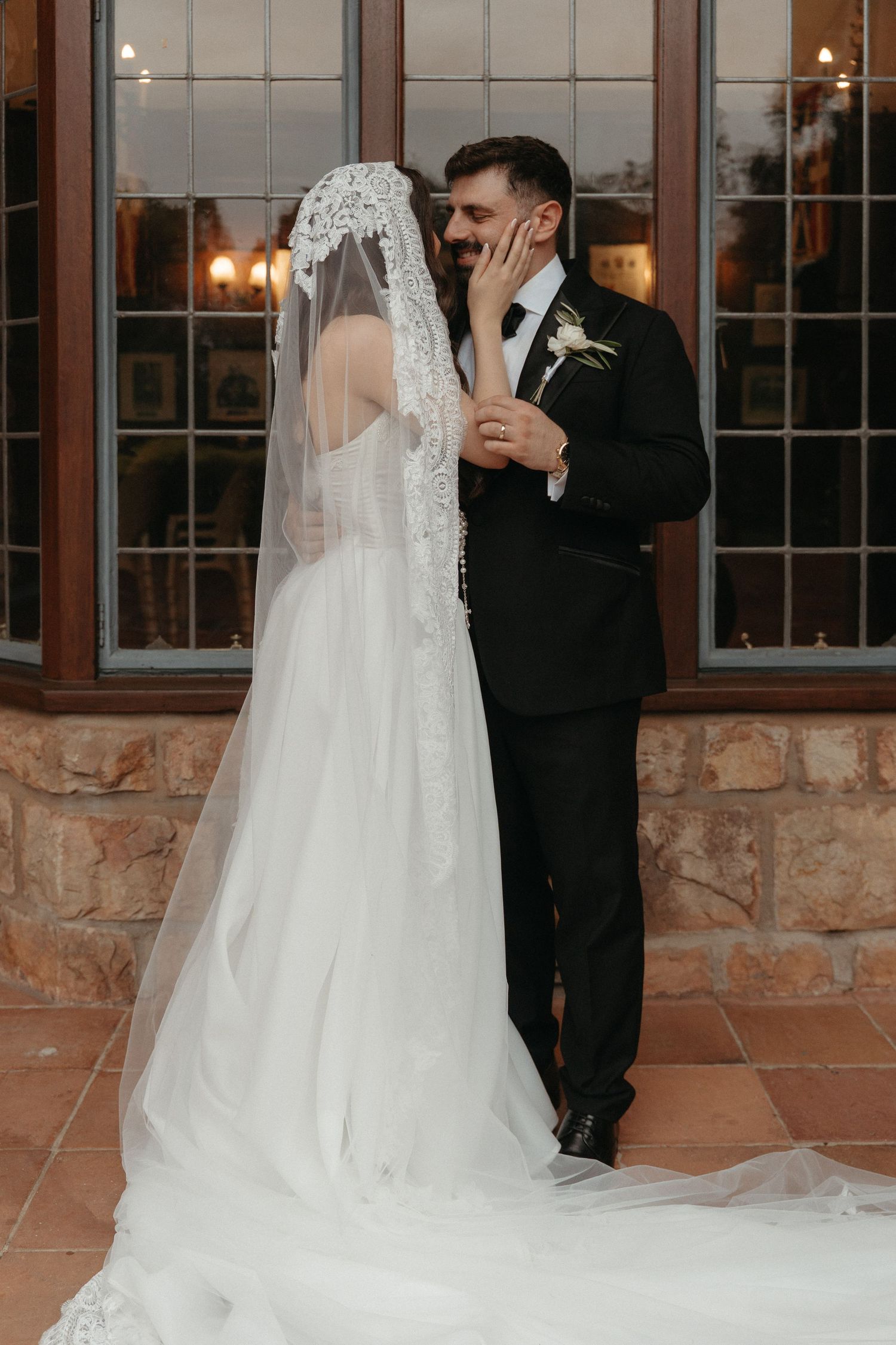 Bride and groom share a romantic kiss, bride in lace veil and white gown, groom in black suit with boutonniere.