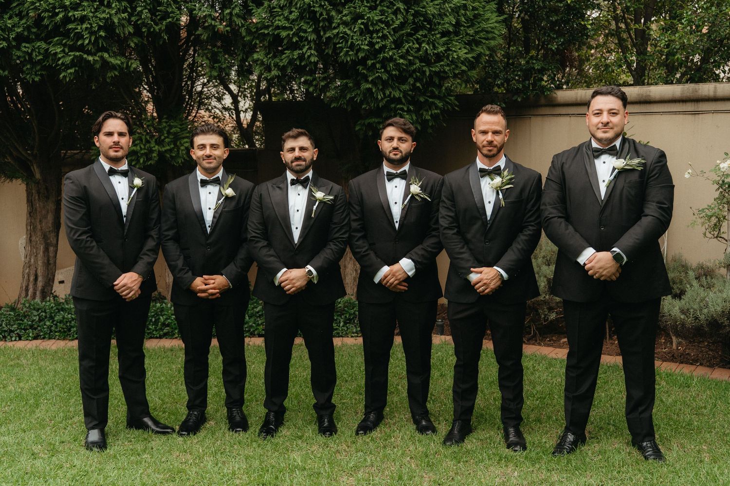 Six groomsmen in black tuxedos and bow ties pose outdoors on green lawn at elegant wedding venue.