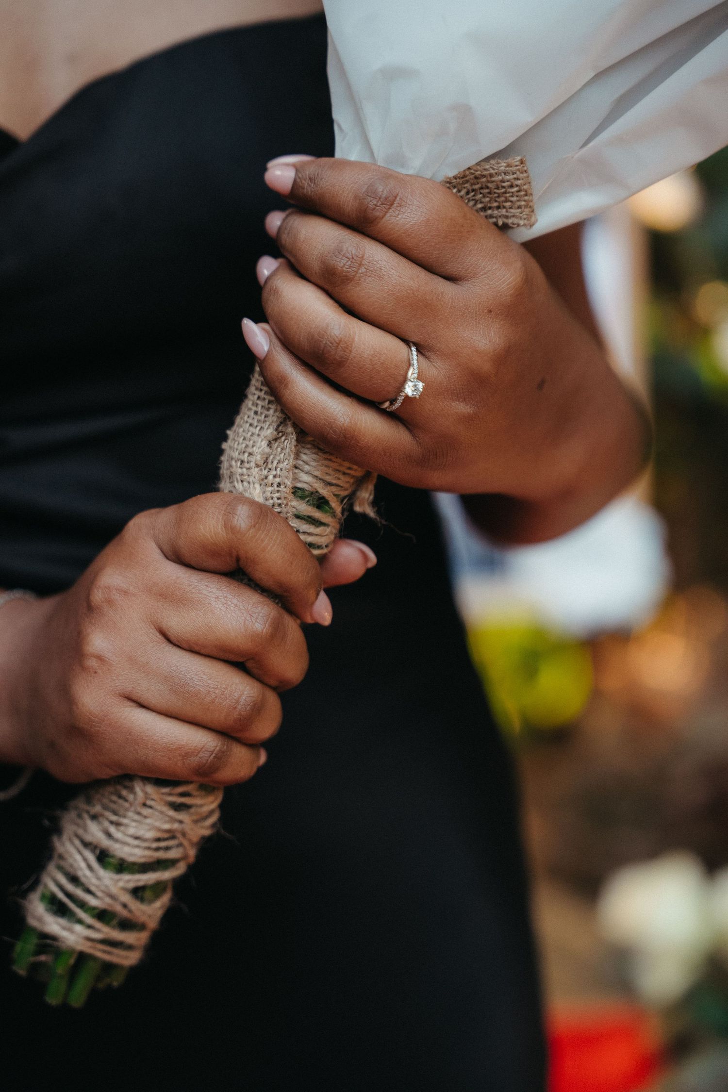 Close-up of bride holding burlap-wrapped bouquet, diamond engagement ring visible on finger, wedding ceremony.