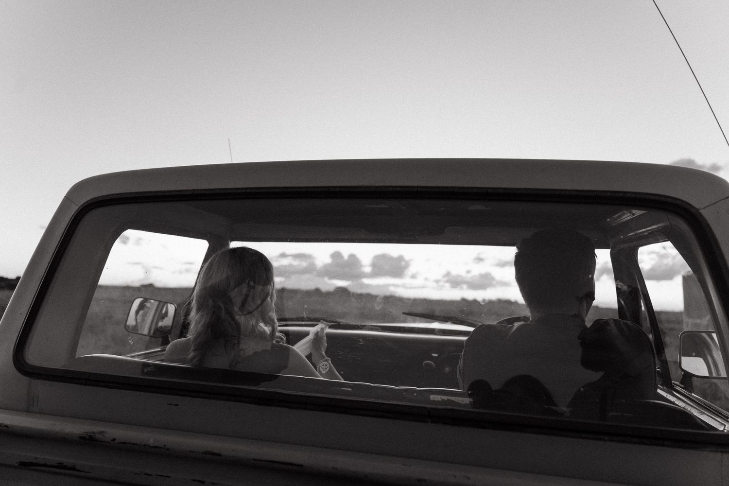 Black and white photo of two people sitting in the front seat of a vintage truck, viewed through the rear window.