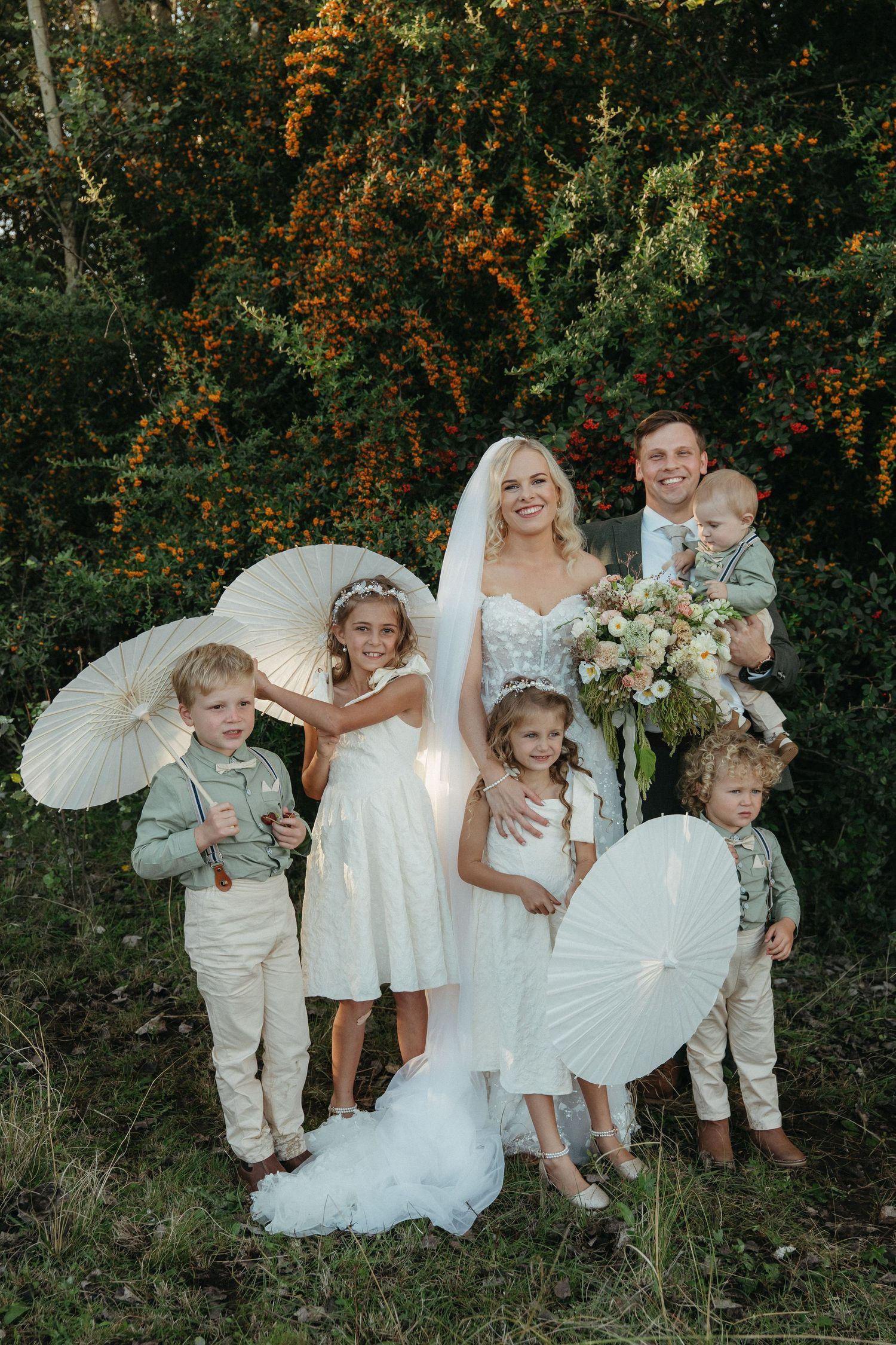 Bride and groom pose with children holding white parasols in front of lush greenery at an outdoor wedding ceremony.