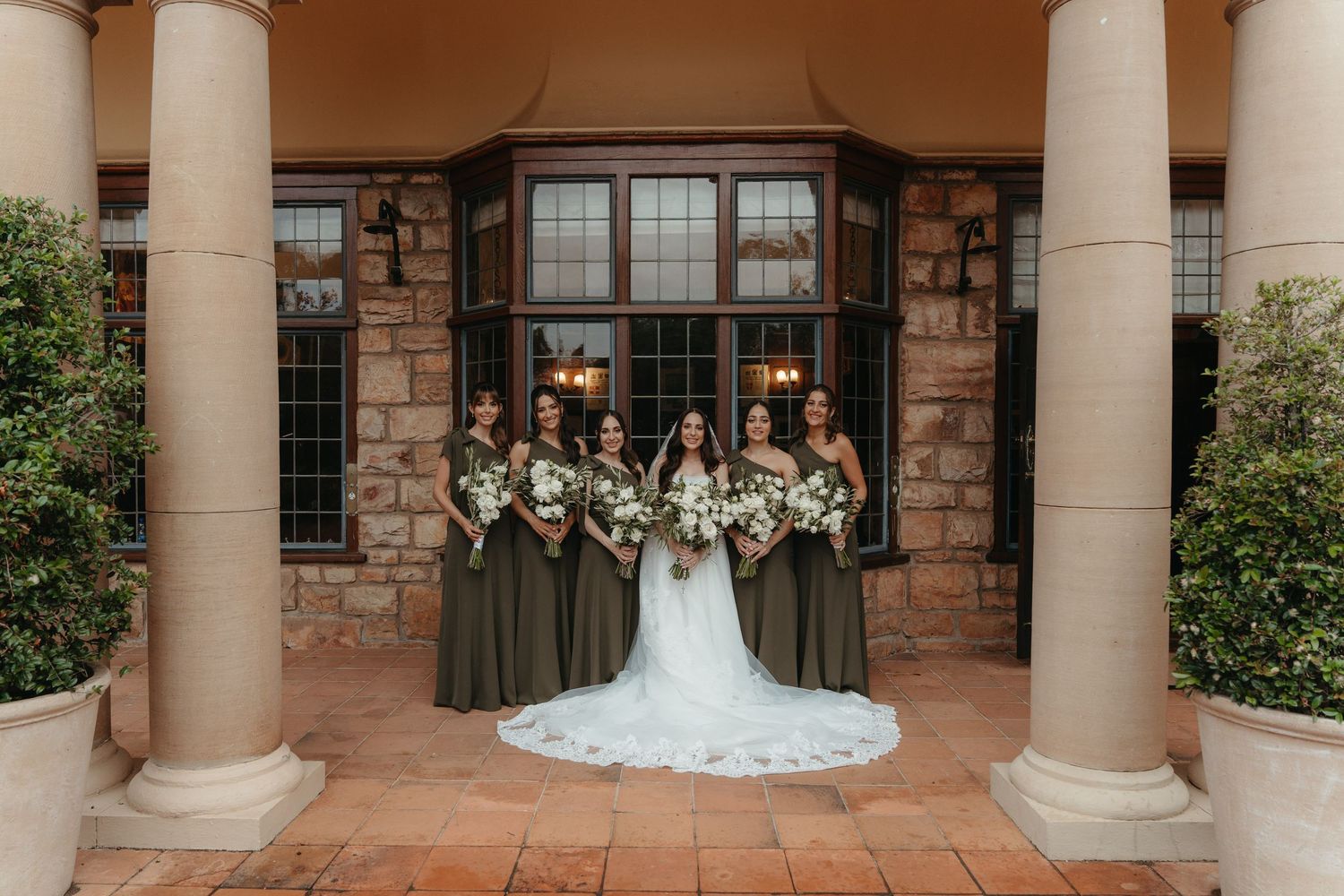Bride and bridesmaids in olive green gowns pose before stone columns of Glenshiel in Johannesburg