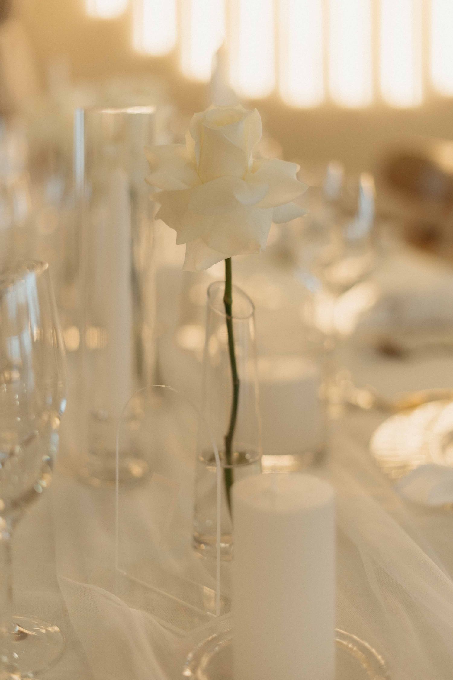 Elegant white rose in glass vase on wedding reception table with candles and glassware.