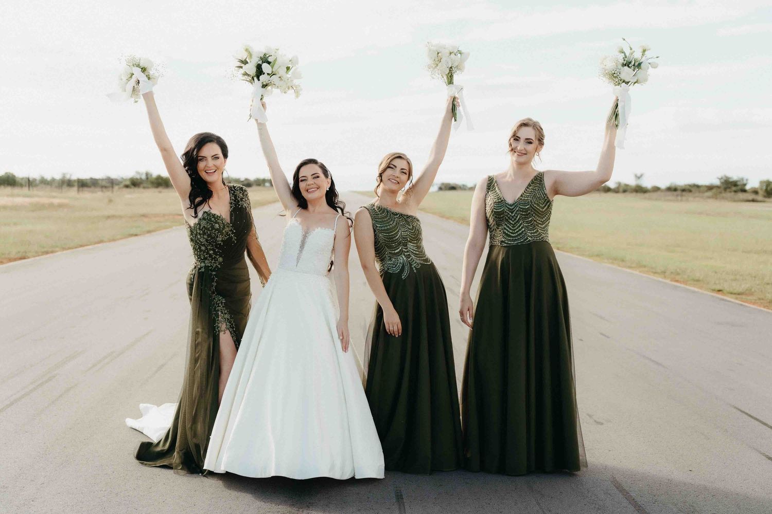 Bride in white gown with three bridesmaids in dark green dresses raising bouquets outdoors.