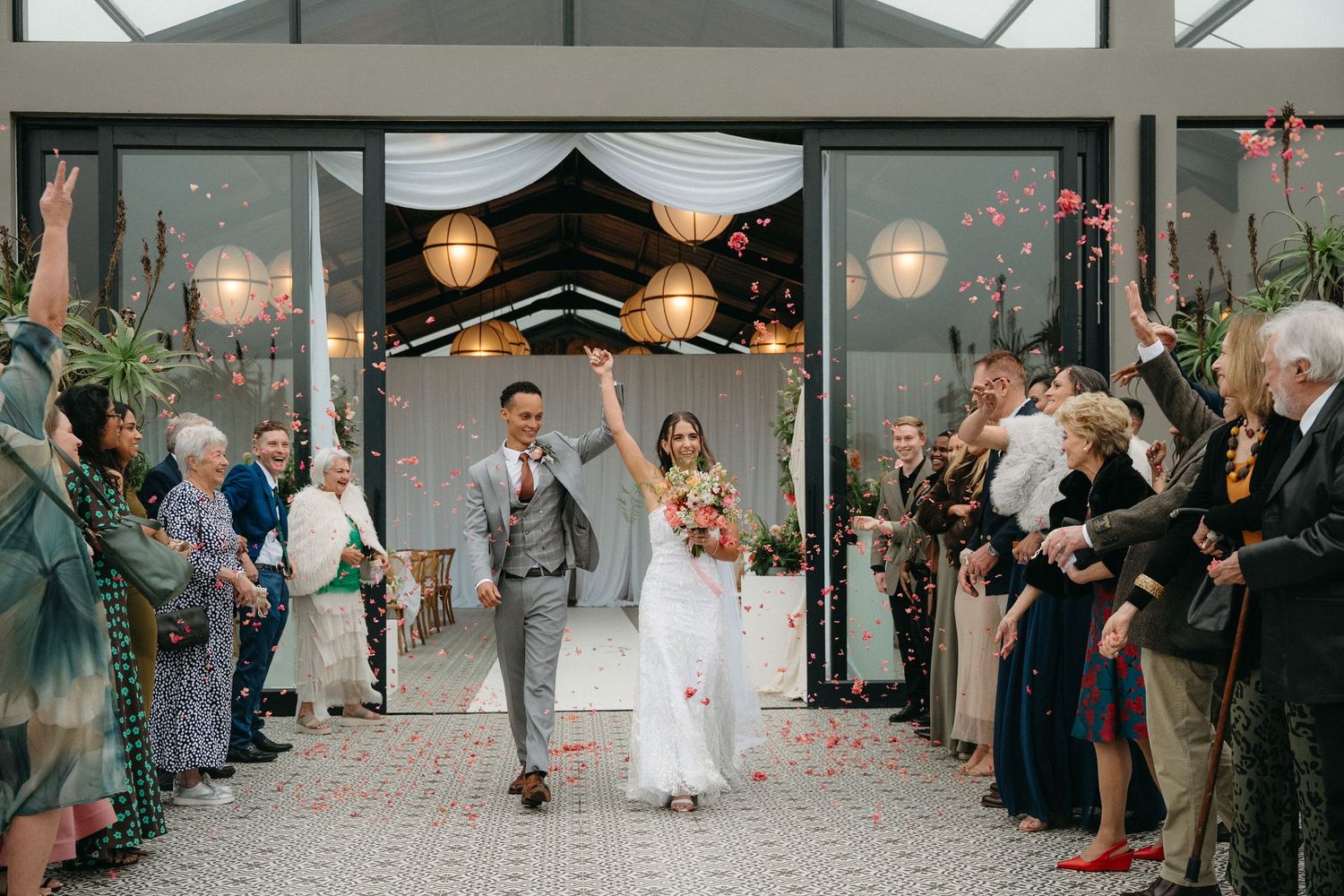 Joyful newlyweds exit wedding ceremony through confetti shower, guests cheering in elegant venue with string lights.