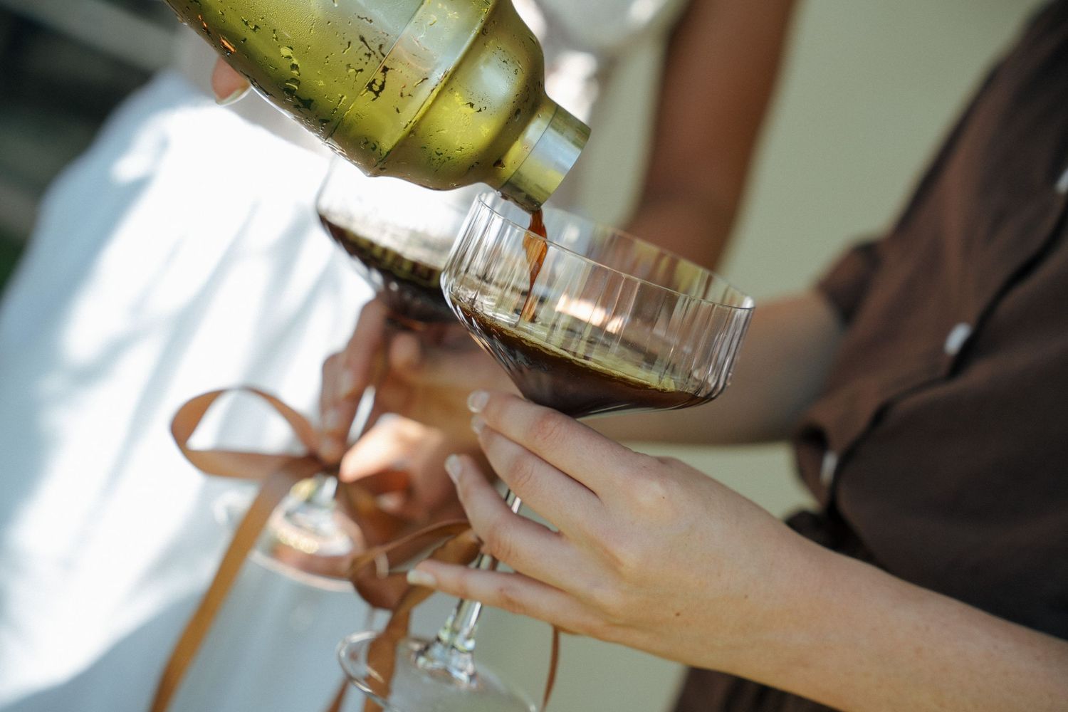 Hands hold wine glasses tied with brown ribbon while espresso martini is poured from a gold cocktail shaker at an outdoor celebration.