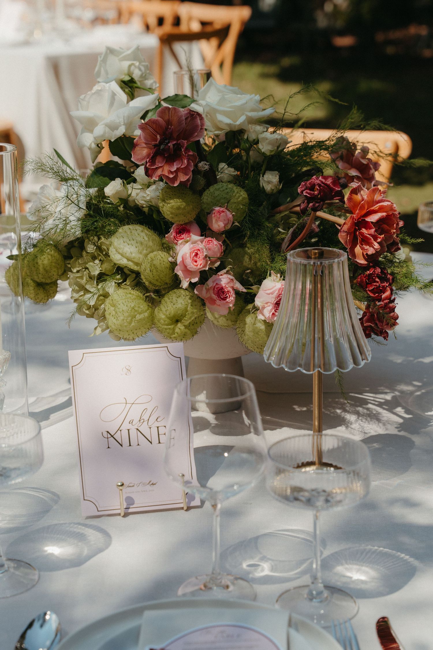 Elegant wedding table nine centerpiece with pink and burgundy flowers, greenery, crystal glasses, and a glass lamp.