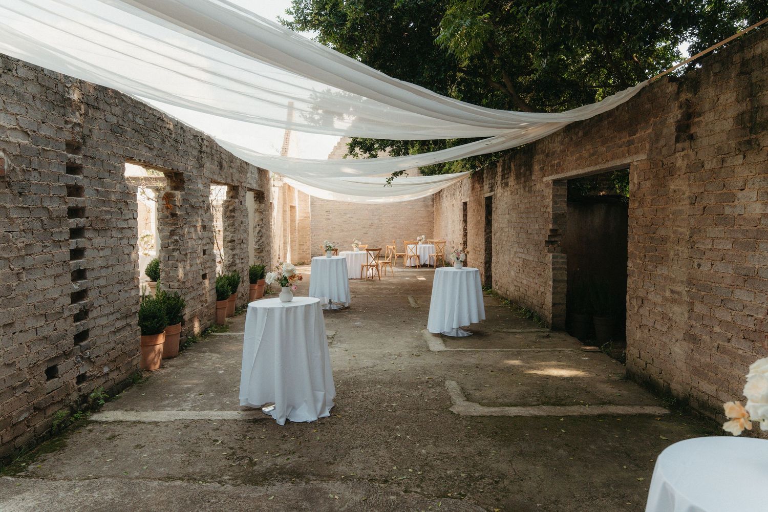 Elegant outdoor wedding reception in rustic stone courtyard with white draped canopy and round tables with white linens.