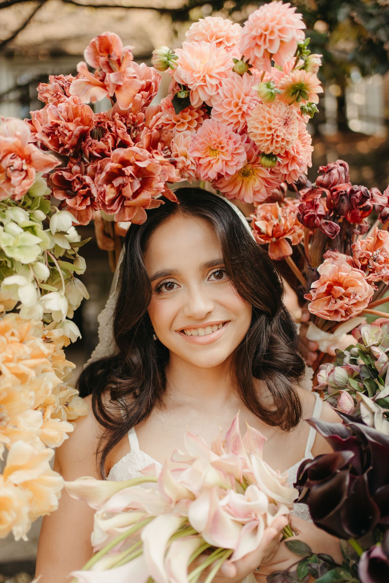 Smiling young woman wearing a floral crown of pink and coral dahlias, holding a bouquet of pastel blooms outdoors.
