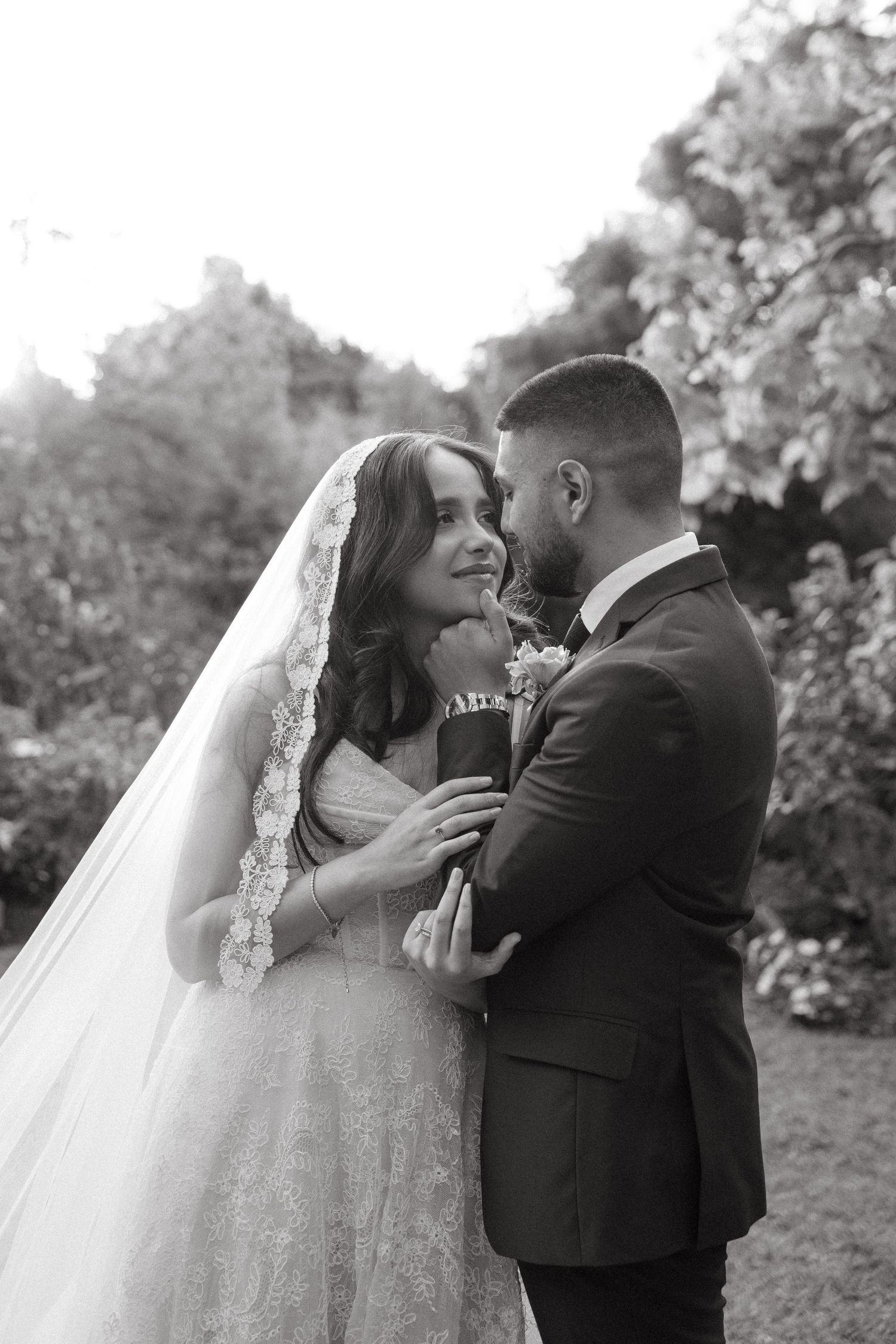 Black and white wedding photo of bride in lace veil and groom in suit embracing outdoors.