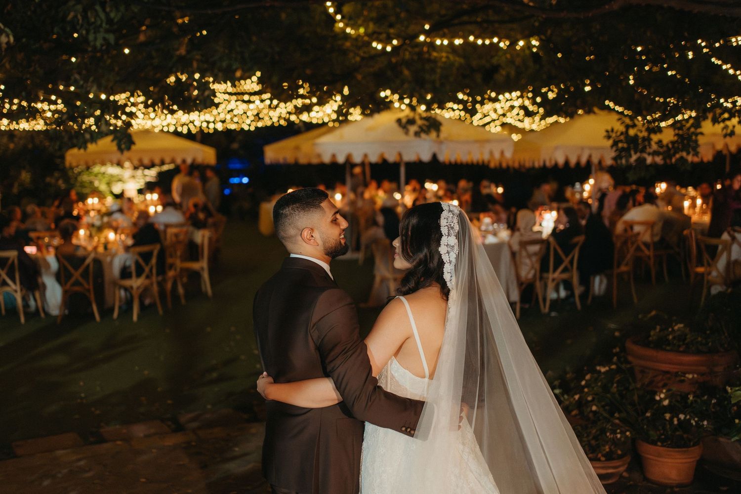 Bride and groom share romantic moment under twinkling string lights at elegant outdoor evening wedding reception.