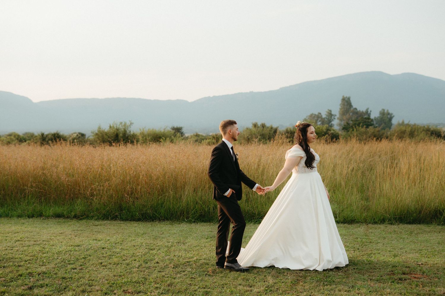 Newlyweds hold hands in golden field with mountain backdrop during sunset photography session.